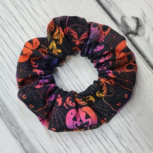 May include: A fabric hair scrunchie featuring a black background with a repeating pattern of orange, pink, and purple gradient jack-o'-lanterns, spiderwebs, and ornate swirling designs. The accessory has a gathered, elasticized construction.