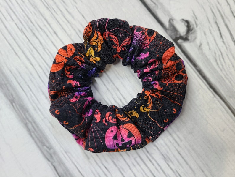 May include: A fabric hair scrunchie featuring a black background with a repeating pattern of orange, pink, and purple gradient jack-o'-lanterns, spiderwebs, and ornate swirling designs. The accessory has a gathered, elasticized construction.