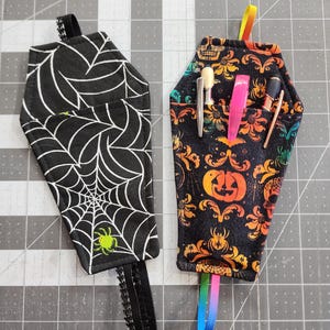May include: Two black fabric pencil holders shaped like coffins. One has a spiderweb pattern and a green spider. The other has a black and orange Halloween pattern with a pumpkin and skulls. Both have black elastic loops.