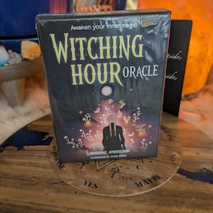 Op de afbeelding: Een rechthoekig kaartspel Witching Hour Oracle in plastic folie. De zwarte doos heeft gele tekst, een centrale illustratie van een persoon met gevlochten haar, paddenstoelen en florale elementen, met de namen van de makers.