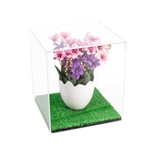 May include: A clear acrylic cube display case containing a white egg-shaped planter with a jagged top edge. Inside the planter are artificial flowers in shades of pink and purple with green leaves. The base is covered in a patch of green artificial grass.