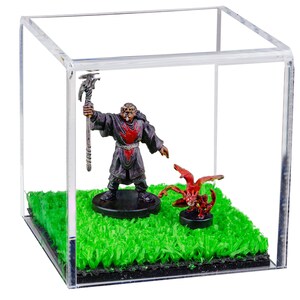 May include: A miniature figure of a red-robed wizard holding a staff stands on a green grass base, facing a red miniature dragon figure with wings spread, also on a green grass base, inside a clear acrylic display cube.