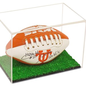 May include: A white and orange football with black laces and a signature on the side. The football is in a clear acrylic display case with a green grass base. The signature reads "Heath Shuler #21 Go Vols".