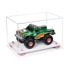 May include: A green toy truck with orange flames and the word "HESS" on the side, displayed in a clear acrylic display case with a white base and pink rubber feet.