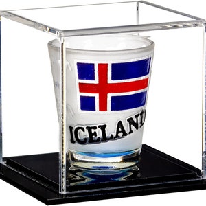 May include: A clear acrylic display case with a black base, showcasing a white shot glass with a blue, red, and white Icelandic flag design and the word "ICELAND" printed on it.