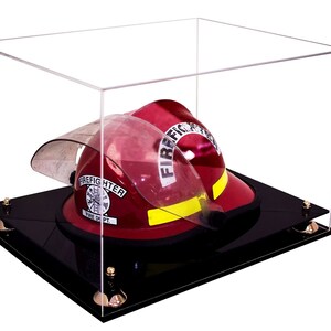 May include: A red firefighter helmet with a black visor and a gold badge that says "Firefighter Fire Dept." is displayed in a clear acrylic display case with a black base.