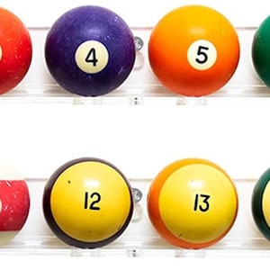 May include: A set of fifteen numbered billiard balls and one white cue ball arranged in two rows on a clear acrylic display rack. The top row features solid colored balls numbered one through eight, while the bottom row displays striped balls nine through fifteen and a plain white ball.