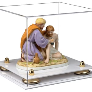 May include: A clear acrylic display case with a white base and gold accents. Inside the case is a porcelain figurine depicting a scene of two figures, one kneeling and the other sitting, with a brown jug on the ground.