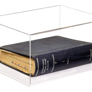 May include: A clear acrylic display case containing a dark blue International Postage Stamp Album Part 1. The book has a textured cover, gold lettering, and aged pages with a small red, white, and blue striped bookmark visible on the side.