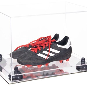 May include: A clear acrylic display case with a black and white soccer cleat with red laces inside. The case has a mirrored base and four black rubber feet.