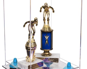 May include: A clear acrylic display case showcasing two gold football trophies. The trophies are mounted on marble bases and blue supports. The case is designed to protect and present awards.