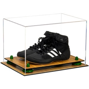 May include: A pair of black wrestling shoes with white stripes are displayed in a clear acrylic display case with a wood base. The case has a wood base with green rubber feet.