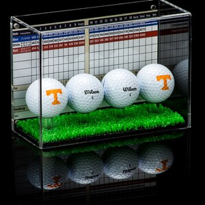 Op de afbeelding: Een transparante acryl display met een groene grasbasis bevat vier golfballen. Drie van de golfballen zijn wit met het woord "Wilson" en het nummer "4" erop gedrukt. De vierde golfbal is wit met een oranje "T" erop gedrukt. De display heeft een gerasterd diagram aan de achterkant met nummers en tekst.