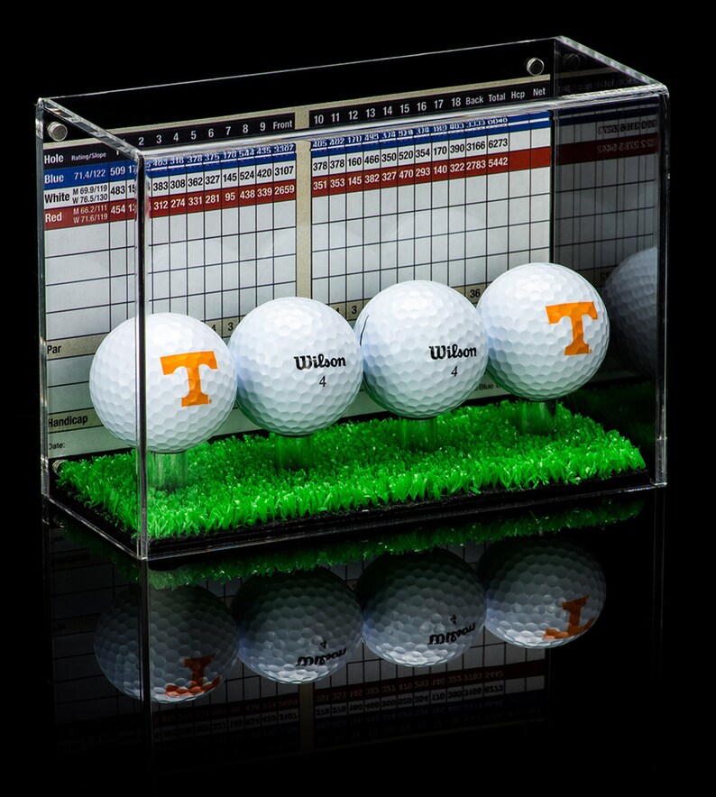 Op de afbeelding: Een transparante acryl display met een groene grasbasis bevat vier golfballen. Drie van de golfballen zijn wit met het woord "Wilson" en het nummer "4" erop gedrukt. De vierde golfbal is wit met een oranje "T" erop gedrukt. De display heeft een gerasterd diagram aan de achterkant met nummers en tekst.