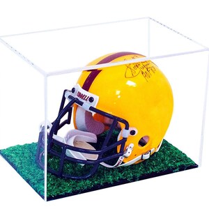 May include: A yellow football helmet with a dark blue face mask and a maroon stripe, featuring a signature on the side. The helmet is displayed inside a clear rectangular acrylic case on a base covered in green artificial turf grass.
