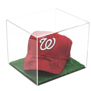 May include: A red baseball cap with a white "W" logo and black signatures on the front, displayed in a clear acrylic display case with a green grass base.