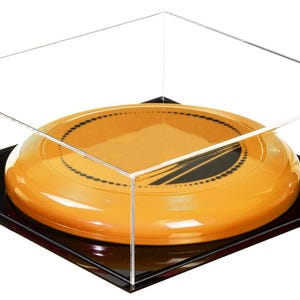 May include: An orange and black frisbee is displayed inside a clear acrylic display case. The frisbee has a black circular design and is set on a black base. The case is square and transparent, showcasing the frisbee.