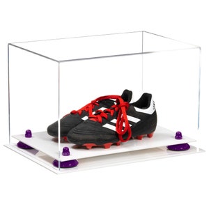 May include: A clear acrylic display case with a white base and purple rubber feet. A pair of black and red soccer cleats are displayed inside the case.