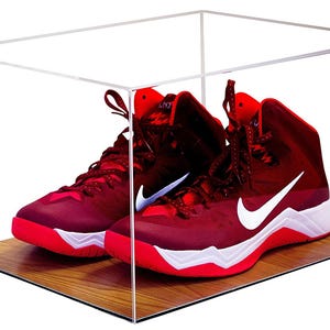 May include: A clear acrylic display case containing a pair of high-top athletic sneakers. The sneakers are primarily dark red with white Nike swoosh logos and bright red soles. The display case features a rectangular wooden base with a grain pattern.