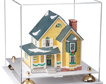 May include: A miniature yellow house with a dark green roof and white trim, featuring snow accents, a brick chimney, and holiday wreaths. The model is enclosed in a clear acrylic display case with a white base and four gold-toned metal feet.