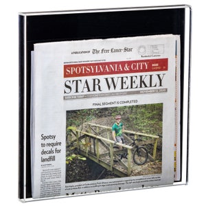 May include: A clear acrylic newspaper holder with a black border, displaying the "Spotsylvania & City Star Weekly" newspaper dated November 12, 2020. The newspaper features articles and a photo of a person on a bicycle. The holder protects and presents the newspaper.