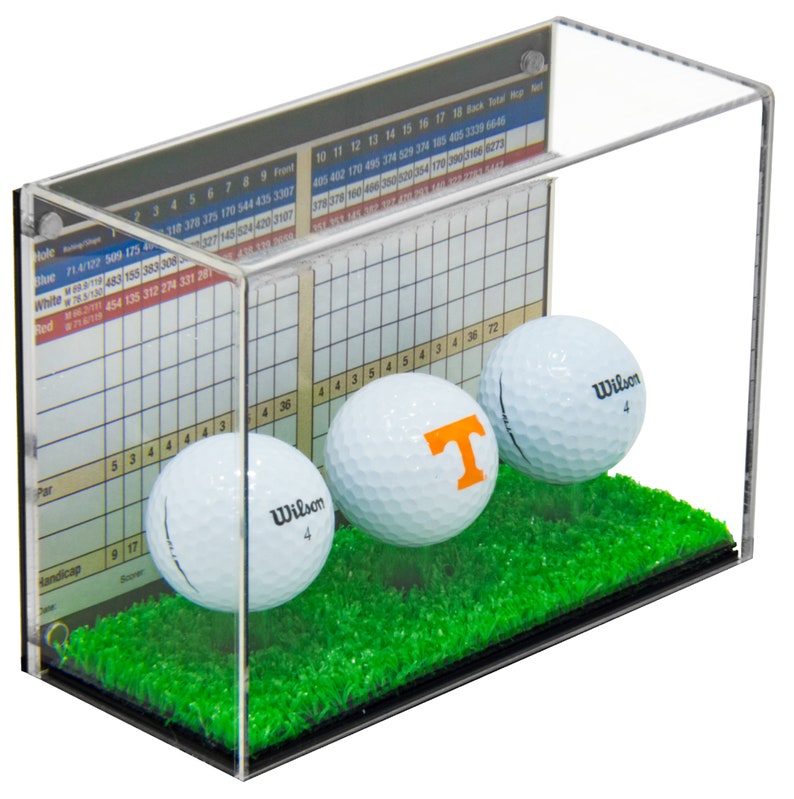 Op de afbeelding: Een transparante acryl displaykast met een groene kunstgrasbasis. Drie witte golfballen worden in de vitrine tentoongesteld. Een golfbal heeft een grote oranje "T" erop. De vitrine heeft een scorekaart aan de zijkant met nummers en tekst.