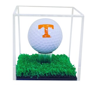 May include: A white golf ball with an orange "T" on it, sitting on a green artificial grass base inside a clear acrylic cube.