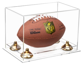 May include: A brown leather Wilson NFL football with white laces, featuring the text THE DUKE and an NFL logo, displayed inside a clear rectangular acrylic case supported by four gold-colored metal risers with rounded bases and nut tops.