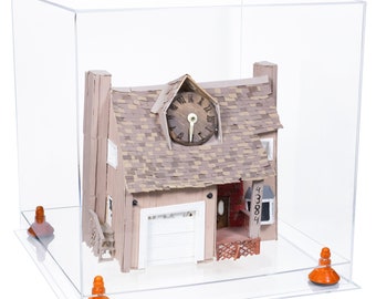 May include: A miniature house model with a clock on the roof, displayed inside a clear acrylic box. The house has a beige exterior, a gray shingled roof, and a white garage door. Orange supports elevate the box.