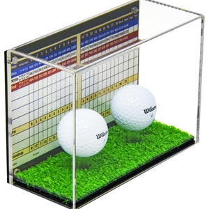 Op de afbeelding: Een transparante acryl display met twee witte golfballen op een groene kunstgrasbasis. De display heeft een scorekaart aan de zijkant met de tekst "Red 71 6/11/19 454 135 312 274 331 281".