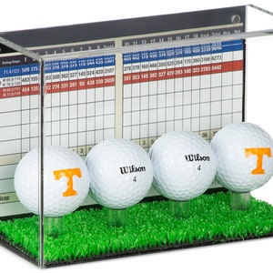 Op de afbeelding: Een transparante acryl displaykast met een groene grasbasis en vier golfballen. De golfballen zijn wit met oranje en zwarte logo's. De kast heeft een scorekaart aan de zijkant met de tekst "Total Men Net".