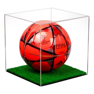 May include: A red and black soccer ball with the text "UEFA CHAMPIONS LEAGUE" and "FINAL MILANO 2016" displayed on the ball, inside a clear acrylic display case with a green artificial grass base.