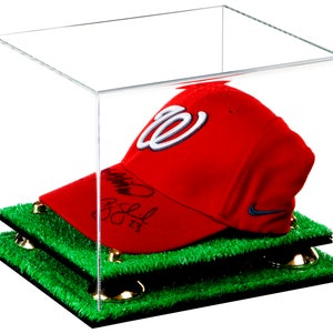 May include: A red baseball cap with a white "W" logo and a signature on the side, displayed in a clear acrylic display case with a green grass base.