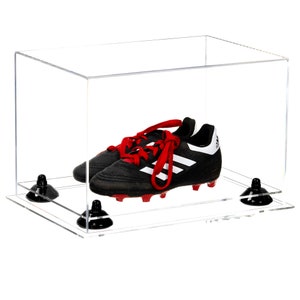 May include: A clear acrylic display case with a pair of black and white soccer cleats with red laces inside. The case has a black base with four suction cups.