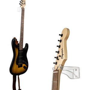 May include: An electric guitar with a black and yellow sunburst finish, hanging on a clear wall mount. The headstock displays the "Squier Stratocaster" logo. A brown leather strap is attached.