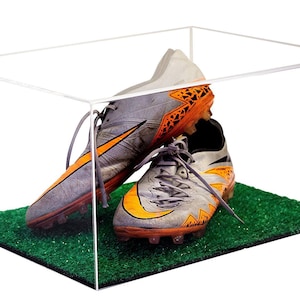 May include: A pair of gray and orange soccer cleats are displayed in a clear acrylic display case with a green artificial grass base.