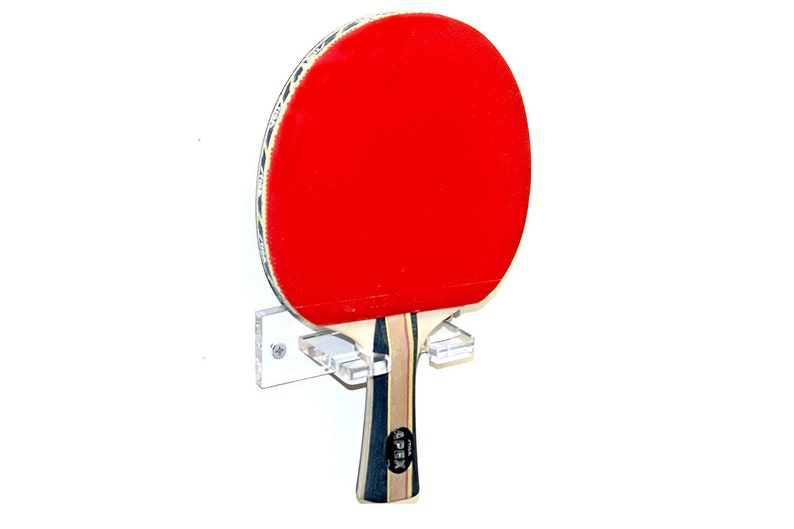 May include: A clear acrylic wall mount for storing two ping pong paddles. The mount is attached to a white wall with screws. A red and black ping pong paddle is mounted on the holder.