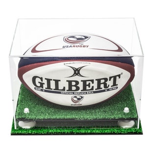 May include: A Gilbert USA Rugby official replica ball in size 5, featuring white, navy, and maroon panels with the USA Rugby logo. The ball is displayed inside a clear acrylic case on a green artificial turf base with four white support pegs.