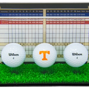Op de afbeelding: Een transparante acryl display met drie golfballen. De golfballen zijn wit met zwarte letters die "Wilson" en het nummer "4" zeggen. Een golfbal heeft een grote oranje "T" erop. De display heeft een scorekaart met nummers en een grasachtige groene basis.