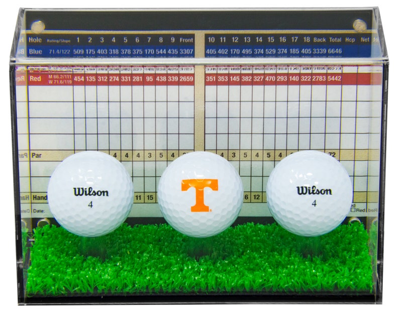 Op de afbeelding: Een transparante acryl display met drie golfballen. De golfballen zijn wit met zwarte letters die "Wilson" en het nummer "4" zeggen. Een golfbal heeft een grote oranje "T" erop. De display heeft een scorekaart met nummers en een grasachtige groene basis.