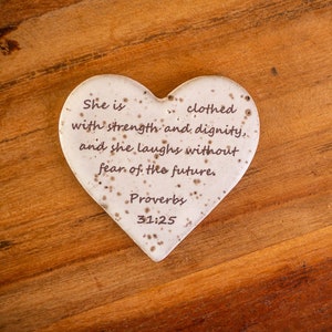 May include: A white heart-shaped ceramic ornament with a brown inscription that reads "She is clothed with strength and dignity, and she laughs without fear of the future. Proverbs 31:25". The ornament is on a wooden surface.