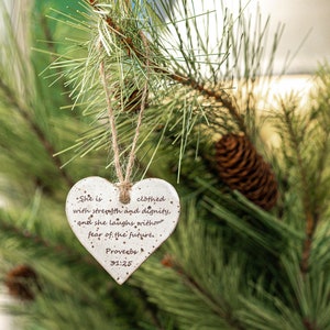 May include: A white heart-shaped ornament with a brown twine hanger. The ornament has a quote from Proverbs 31:25 inscribed on it: "She is clothed with strength and dignity, and she laughs without fear of the future."