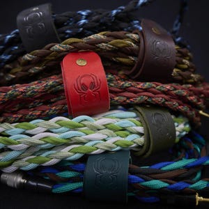 Pode incluir: Uma coleção de cabos trançados em várias cores, incluindo azul, castanho, verde e vermelho, cada um preso com uma tira de couro com um logótipo de aranha em relevo e um pequeno rebite metálico. Os cabos têm conectores metálicos nas pontas.