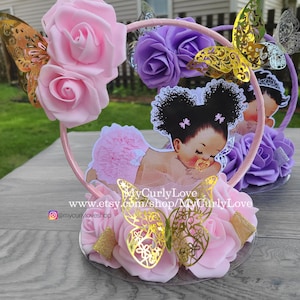 Pode incluir: Um centro de mesa circular rosa com um recorte de um beb&eacute; de cabelo escuro com marias-chiquinhas, vestindo um tutu rosa e uma chupeta. O aro &eacute; decorado com rosas de espuma rosa e roxas, borboletas douradas e o texto MyCurlyLove.