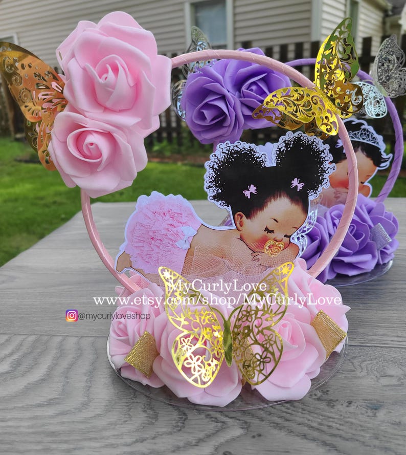 Pode incluir: Um centro de mesa circular rosa com um recorte de um beb&eacute; de cabelo escuro com marias-chiquinhas, vestindo um tutu rosa e uma chupeta. O aro &eacute; decorado com rosas de espuma rosa e roxas, borboletas douradas e o texto MyCurlyLove.