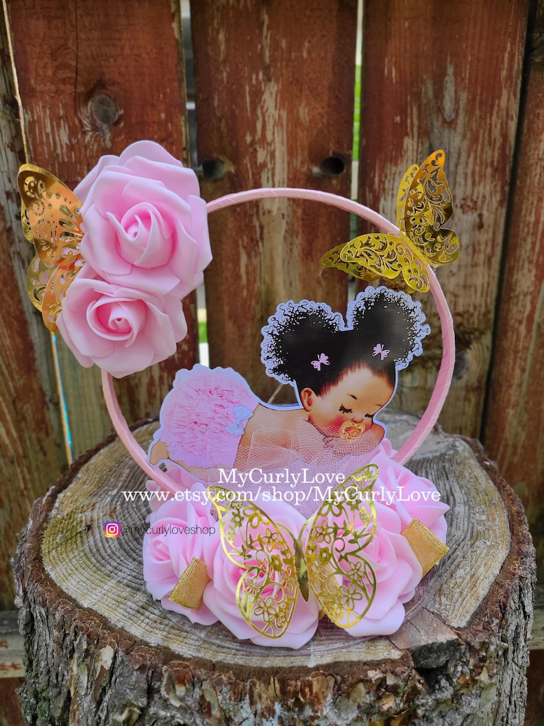 Pode incluir: Um topo de bolo circular rosa com duas rosas de espuma, tr&ecirc;s borboletas de filigrana met&aacute;lica dourada e um recorte central de um beb&eacute; com cabelo encaracolado escuro, tutu rosa e chupeta, com o texto MyCurlyLove www.etsy.com/shop/MyCurlyLove.