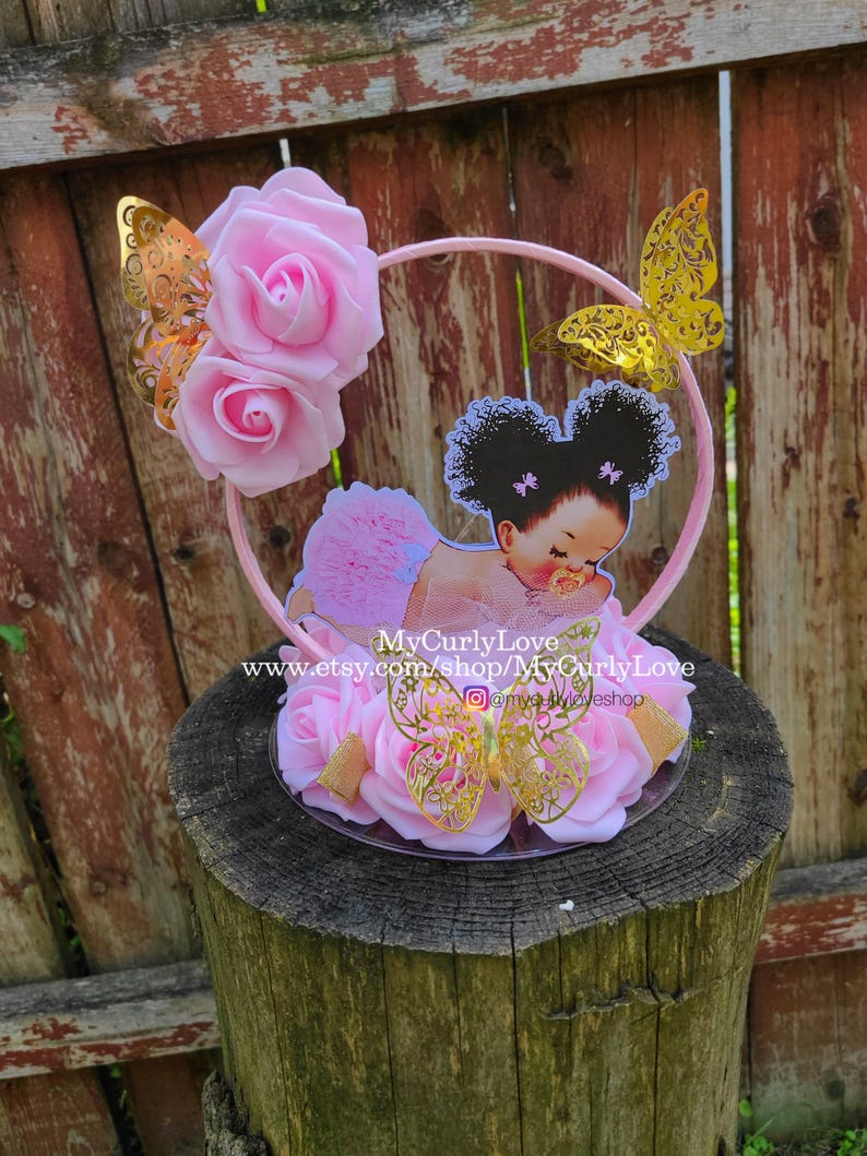 Pode incluir: Um topo de bolo com uma moldura circular rosa decorada com rosas de espuma rosa claro e tr&ecirc;s borboletas douradas met&aacute;licas. Um recorte de um beb&eacute; com cabelo encaracolado escuro e roupa rosa est&aacute; centrado, com o texto MyCurlyLove.