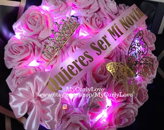 May include: A bouquet of glittery pink artificial roses illuminated by internal lights, featuring a gold-toned tiara, a pink ribbon with the text "Quieres Ser Mi Novia" in gold lettering, a gold butterfly cutout, and a pink satin bow.
