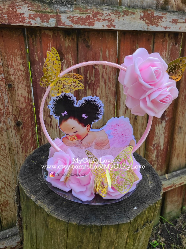 Pode incluir: Um centro de mesa decorativo com uma ilustra&ccedil;&atilde;o de beb&eacute; com cabelo escuro encaracolado e uma chupeta, rodeado por rosas de espuma cor-de-rosa e recortes de borboletas met&aacute;licas douradas numa moldura circular. Texto: MyCurlyLove www.etsy.com/shop/MyCurlyLove.
