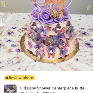 Pode incluir: Um centro de mesa decorativo de v&aacute;rios n&iacute;veis com rosas de espuma roxas, recortes de borboletas com glitter dourado e um recorte central de um beb&eacute; com uma tiara. A estrutura &eacute; adornada com numerosas pequenas borboletas roxas e cor-de-rosa.